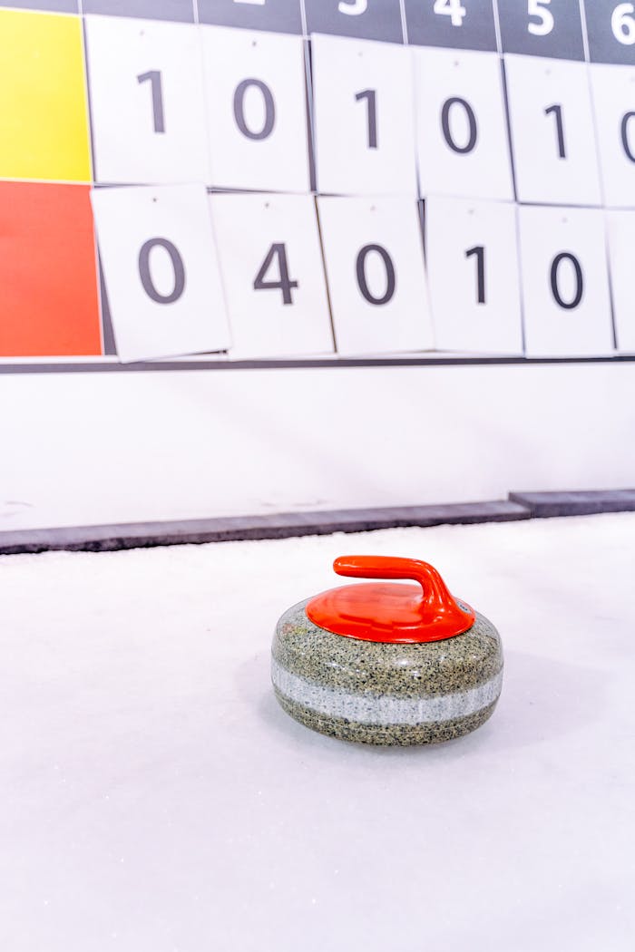 Close-up of a curling stone with a red handle on an ice rink, scoreboard in the background.